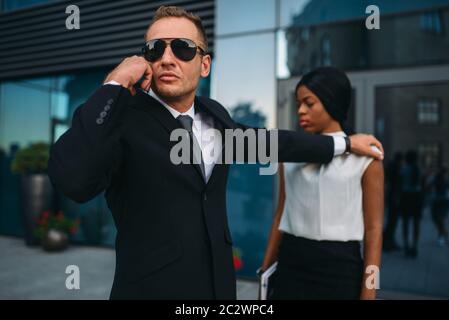 Ernsthafte Leibwächter in Anzug und Sonnenbrille Zugriffe auf Hörer für weibliche client Schutz. Security Guard ist eine riskante Beruf, professionelle Stockfoto
