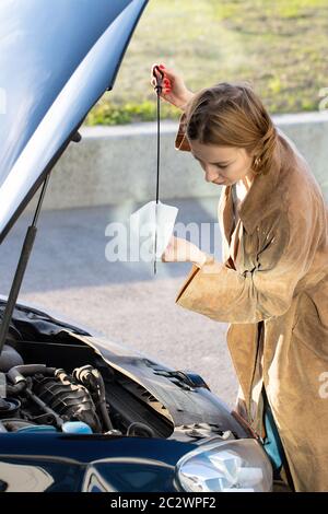 Selbstbewusste kaukasische Frau Fahrer Überprüfung Ölstand im Auto Motor Messstab, Stadtbild auf Hintergrund. Stockfoto