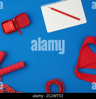 Rotes Sportshirt, rotes Springseil und kabellose Kopfhörer in roter Hülle auf blauem Grund, Draufsicht, leeres weißes Blatt Papier, flaches Lay Stockfoto