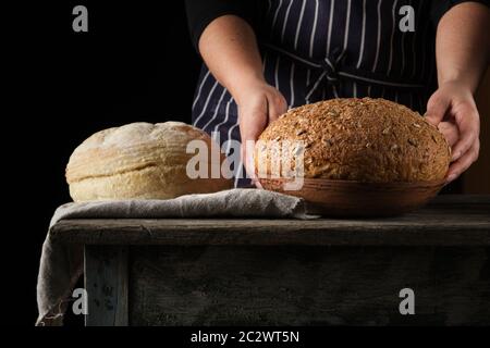 Frau im blau gestreiften Schürze hält in ihrer Hand gebackene runde Roggenbrot und mit Sonnenblumenkernen, hölzernen Tisch Stockfoto