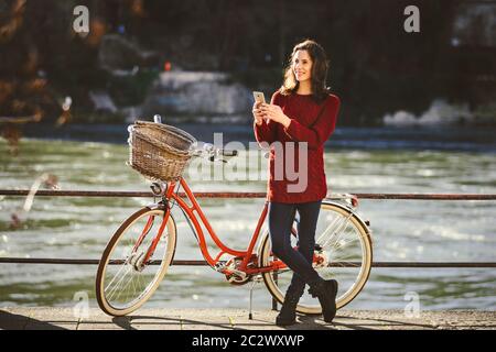 Thementourismus mit Fahrrad und moderner Technik. Schöne junge kaukasische Frau steht in der Nähe des roten Retro-Fahrrads am Fluss am Fluss Stockfoto