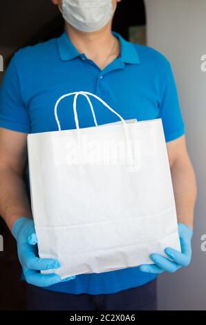 Lieferer hält einen Papierbeutel mit Lebensmitteln in Gummihandschuhen und einer Schutzmaske. Schnelle und kostenlose Lieferung der Ware an die Tür. Online-Shop und Stockfoto