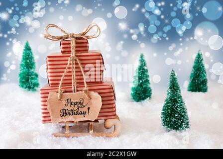 Schlitten mit Weihnachtsgeschenk und Label mit englischen Kalligraphie Happy Birthday. Schnee mit Weihnachtsbaum und Blau schimmernden Hintergrund Stockfoto