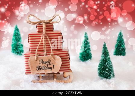 Schlitten mit Weihnachtsgeschenk und Label mit englischen Kalligraphie Happy Birthday. Schnee mit Weihnachtsbaum und Rot funkelnden Hintergrund Stockfoto