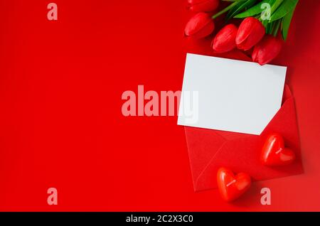 Grußkarte mit Liebe. Roter Umschlag mit leerem Papier, Blumenstrauß aus Tulpen und herzförmigen Kerzen. Draufsicht mit Platz für Ihren Text. Flaches Lay Stockfoto