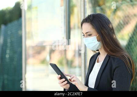 Frau, die eine Schutzmaske trägt, mit Handy in einer Bushaltestelle sitzt Stockfoto