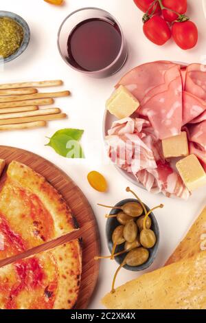 Italienisches Essen. Pizza, Käse, Schinken, Kapern, Pesto, Tomaten und Rotwein, geschossen von oben auf weißem Hintergrund Stockfoto