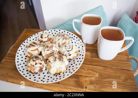 Zwei Tassen schwarzen Tee, frische und duftende Zimtrollen aus nächster Nähe auf einem Teller mit Tupfen stehen auf einem Holztablett. Schöner Morgen. Stockfoto