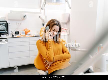Junge Frau in orangefarbenem Pullover spricht am Telefon, während sie darauf wartet, vom Zahnarzt gesehen zu werden Stockfoto
