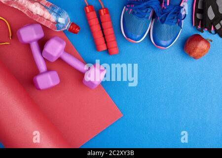 Zwei blaue Turnschuhe mit Schnürsenkeln, einer Flasche Wasser und einer rot verdrehten Neopren-Matte auf blauem Texturhintergrund, Draufsicht, Sportset Stockfoto
