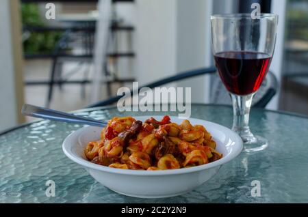 Nahaufnahme einer weißen Schüssel gefüllter Pasta ( Ravioli ) mit Tomatensauce und Pilzen mit einem Glas Rotwein auf einem Balkon Tisch im Freien Stockfoto