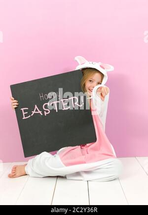 Hübsches blondes Mädchen mit gemütlichem Kaninchen-Kostüm und Tafel mit den Worten Happy easter posiert im Studio und ist glücklich Stockfoto