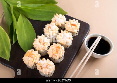 Brötchen mit gelber Sahne-Sauce und gebratenen Garnelen gekrönt. Sushi-Rollen auf dunklem Holzbrett. Beige Hintergrund mit Blättern Stockfoto