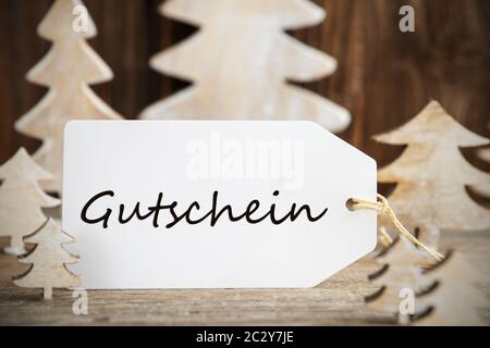 Etikett mit deutschem Text Gutschein Gutschein bedeutet. Weiß Holz- Weihnachten Baum als Dekoration. Braun Holz- Hintergrund Stockfoto