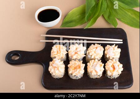 Brötchen mit Sahne-Sauce und gebratenen Garnelen gekrönt. Sushi-Rollen auf dunklem Holzbrett. Beige Hintergrund mit Blättern. Draufsicht Stockfoto