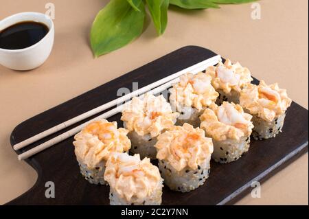 Brötchen mit gelber Sahne-Sauce und gebratenen Garnelen gekrönt. Sushi-Rollen auf dunklem Holzbrett. Beige Hintergrund mit Blättern Stockfoto