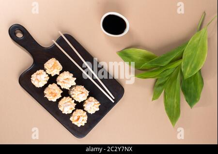 Brötchen mit Sahne-Sauce und gebratenen Garnelen gekrönt. Sushi-Rollen auf dunklem Holzbrett. Beige Hintergrund mit Blättern. Draufsicht Stockfoto