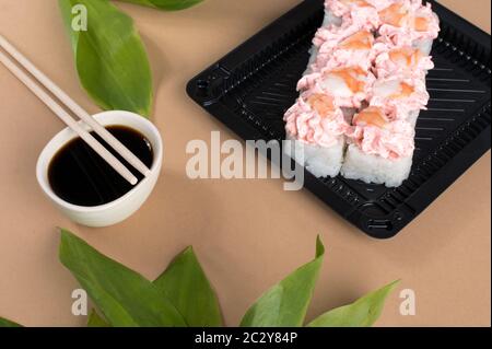 Sushi-Brötchen mit rosa Frischkäse und Garnelen gekrönt. Sushi-Rollen in schwarzen Plastikbehältern gesetzt. Asiatische Lebensmittel Lieferung nach Hause. Beigefarbener Hintergrund Stockfoto