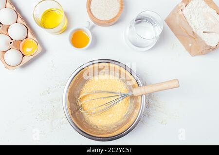 Draufsicht Teig in Metallschüssel mit Schneebesen neben Zutaten zum Kochen hausgemachtes Gebäck mit Mehl, Eiern, Olivenöl und Wasser auf weißem Tisch. Stockfoto