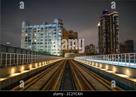 Die Bahngleise des Tramsystems Kaohsiung, das nachts in einen Tunnel mit hohen Gebäuden und Wolkenkratzern im Hintergrund führt Stockfoto