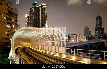 Die Bahngleise des Tramsystems Kaohsiung, das nachts in einen Tunnel mit hohen Gebäuden und Wolkenkratzern im Hintergrund führt Stockfoto