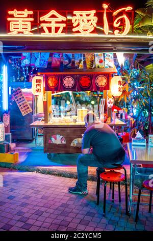 Ein Mann, der sein Smartphone vor einem sehr bunten, neonbeleuchteten kleinen Straßenhändler in der Nacht in Kaohsiung, Taiwan, benutzt Stockfoto