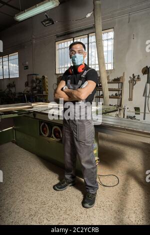 Arbeiter, der mit gekreuzten Armen steht, trägt Gesichtsmaske und Schutzkleidung und blickt in einer Fabrik lächelnd auf die Kamera. Langer Schuss, voller Körper. Stockfoto