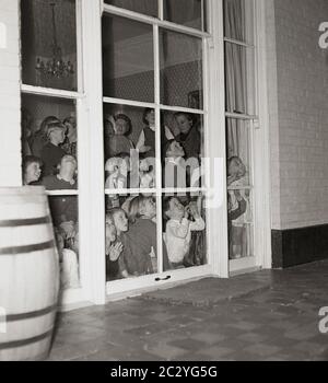 1960er, historisch, ein Grouo von aufgeregt kleinen Kindern auf einer Party in einem Haus spähtend aus den Fenstern auf der Rückseite des Hauses auf die Terrasse draußen, um zu versuchen, und sehen Sie die Überraschung, England, Großbritannien. Stockfoto