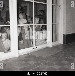 1960er, historisch, ein Grouo von aufgeregt kleinen Kindern auf einer Party in einem Haus spähtend aus den Fenstern auf der Rückseite des Hauses auf die Terrasse draußen, um zu versuchen, und sehen Sie die Überraschung, England, Großbritannien. Stockfoto