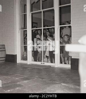 1960er, historisch, ein Grouo von aufgeregt kleinen Kindern auf einer Party in einem Haus spähtend aus den Fenstern auf der Rückseite des Hauses auf die Terrasse draußen, um zu versuchen, und sehen Sie die Überraschung, England, Großbritannien. Stockfoto