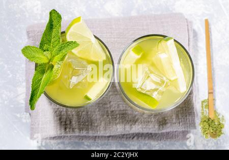 Zwei Gläser eisgrüner Matcha-Tee mit Limette, Eis, frischer Minze und Holzlöffel mit Matcha-Pulver auf grauer Serviette auf hellgrauer Kulisse. Gesunde Ref Stockfoto