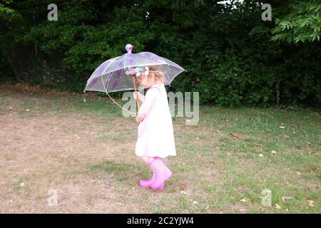 Junge 3-jährige Mädchen mit einem Regenschirm spielen und tragen wellington Stiefel und gefälschte Blumendekoration in den Haaren Stockfoto