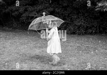 Junge 3-jährige Mädchen mit einem Regenschirm spielen und tragen wellington Stiefel und gefälschte Blumendekoration in den Haaren Stockfoto