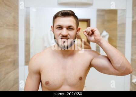 Der Mann reinigt seine Ohren mit Wattestäbchen, morgendlichen routinemäßigen Hygieneverfahren. Athletischer Mann am Spiegel im Bad Stockfoto