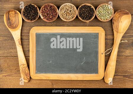 Fünf kleine Holzschüsseln mit verschiedenen Pfefferarten, zwei Kochlöffeln aus olivem Holz und einem alten schwarzen Schieferbrett mit Kopierraum auf einem rustikalen Stockfoto