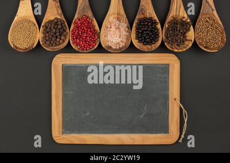 Sieben Kochlöffel aus olivem Holz mit verschiedenen Gewürzen liegen in einer Reihe und eine alte schwarze Schiefertafel mit Kopierraum auf schwarzem Hintergrund Stockfoto