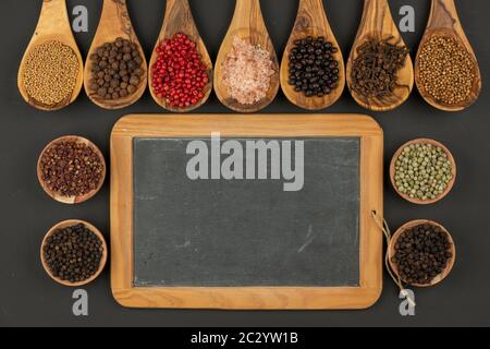 Sieben Kochlöffel aus olivem Holz und vier kleine Holzschüsseln mit verschiedenen Gewürzen und einer alten schwarzen Schiefertafel mit Kopierraum auf einem Schwarz Stockfoto