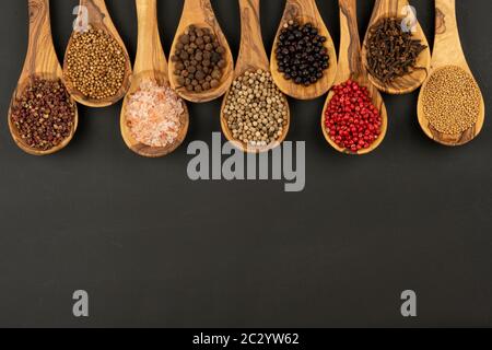 Gewürz und Essen - Neun Kochlöffel aus olivem Holz in einer Reihe mit Gewürz- und Pfeffersorten auf schwarzem Hintergrund mit Kopierraum Stockfoto