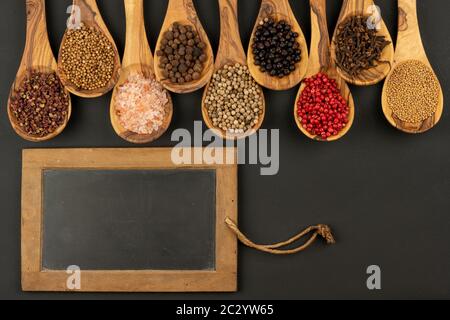 Gewürz und Essen - Neun Kochlöffel aus olivem Holz in einer Reihe mit Gewürz- und Pfeffersorten und einem alten schwarzen Schiefertafel mit Kopierraum Stockfoto