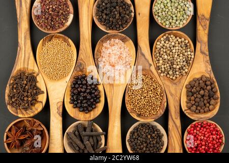 Gewürz und Essen - Hintergrund aus vielen verschiedenen Gewürzen und Pfeffersorten in kleinen Holzschüsseln und Kochlöffeln aus olivem Holz auf einer schwarzen BA Stockfoto