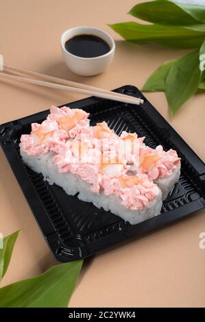 Sushi-Brötchen mit rosa Frischkäse und Garnelen gekrönt. Sushi-Brötchen in schwarzen Plastikbehältern mit Sojasauce und Essstäbchen Stockfoto