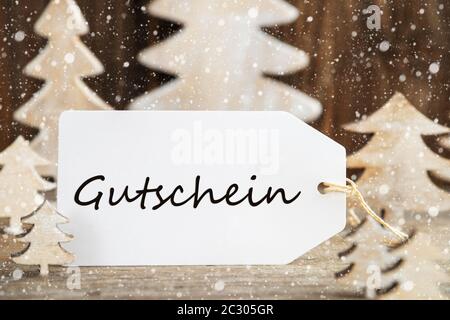 Etikett mit deutschem Text Gutschein Gutschein bedeutet. Weiß Holz- Weihnachten Baum als Dekoration. Braun Holz- Hintergrund mit Schnee Stockfoto