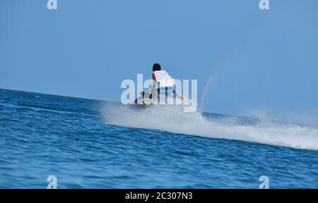 Ein junger Mann, der mit einem Jet-Skier über blauem Meerwasser mit Splash-Spur hinter der Rückansicht mit niedrigem Winkel fährt Stockfoto