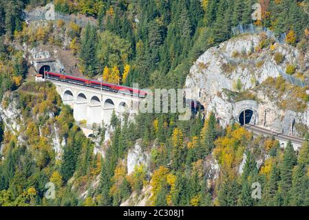 Roter Zug auf einem Viadukt zwischen zwei Tunneln der Semmeringbahn. Die Semmeringbahn ist die älteste Bergbahn Europas und gehört zur UNESCO-Welt Stockfoto