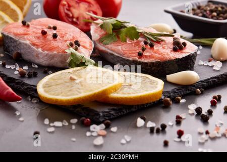 Frisches rohes Lachssteak mit Gewürzen und Gemüse auf einem steinernen Schneidebrett auf schwarzem Hintergrund. Meeresfrüchte-Menü Stockfoto