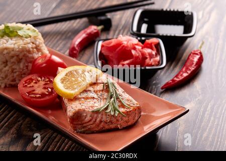 Gegrilltes Lachsfilet mit Reis, eingelegtem Ingwer, Sojasauce, Tomaten, Zitrone und Kräutern auf dunklem Holzhintergrund. Gesunde Ernährung. Stockfoto