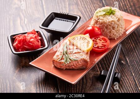 Gegrilltes Lachsfilet mit Reis, eingelegtem Ingwer, Sojasauce, Tomaten, Zitrone und Kräutern auf dunklem Holzhintergrund. Gesunde Ernährung. Stockfoto