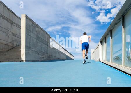 Junger Läufer in blauer Hose und weißem T-Shirt läuft in der Sonne auf blauer Bahn. Motivationskonzept, Karriere Stockfoto