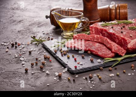 Stillleben mit rohem Fleisch Rinderfilet auf schwarzem Stein Schneidebrett, Rosmarin Zweige, Set aus verschiedenen Paprika, Salz und Olivenöl auf dunklem Hintergrund. F Stockfoto