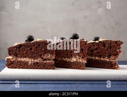 Drei Dreiecksstücke Schokoladenbiskuitkuchen mit Buttercreme auf weißem Holzbrett, grauer Hintergrund Stockfoto